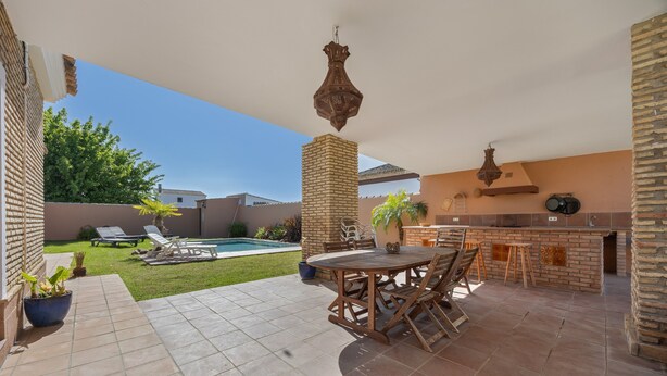 An outdoor patio with a pool, a wooden table and chairs, a brick column, and a hanging light fixture.