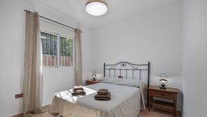 A bedroom with a bed, two bedside tables with lamps, a window with curtains, and a tiled floor.
