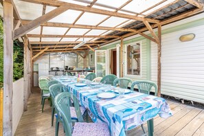Un área de comedor al aire libre cubierta con una mesa lista para cuatro personas, rodeada de sillas verdes, bajo una estructura de vigas de madera.