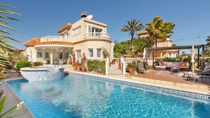 Une piscine avec un bord carrelé, un espace salon couvert et un bâtiment doté d’un balcon.