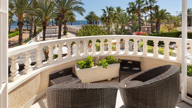 Un balcon agrémenté de meubles en osier, d’un jardinier avec des plantes vertes et offrant une vue sur les palmiers et la mer.
