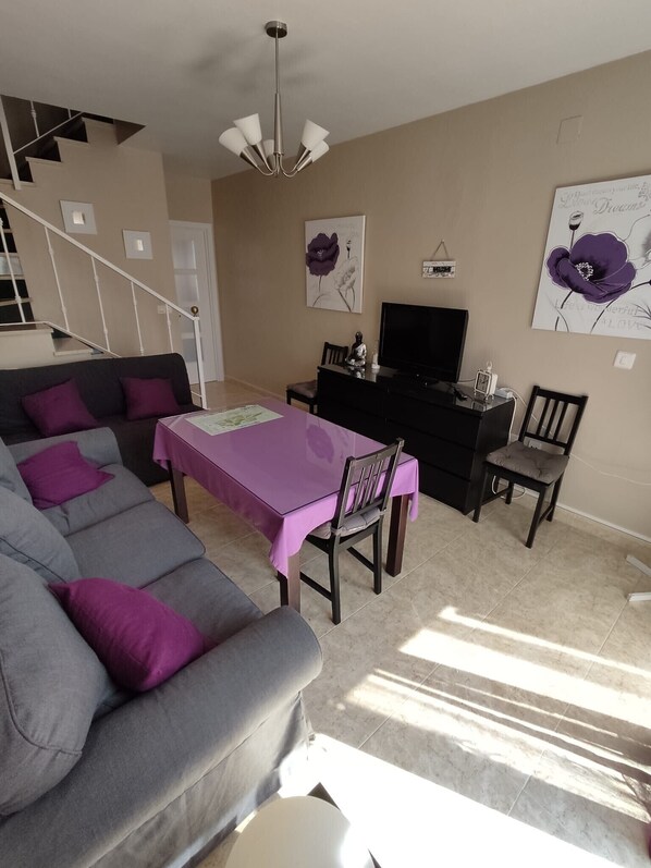 A living room with a gray sofa, purple cushions, a dining table with a purple tablecloth, a black TV stand, and a staircase.