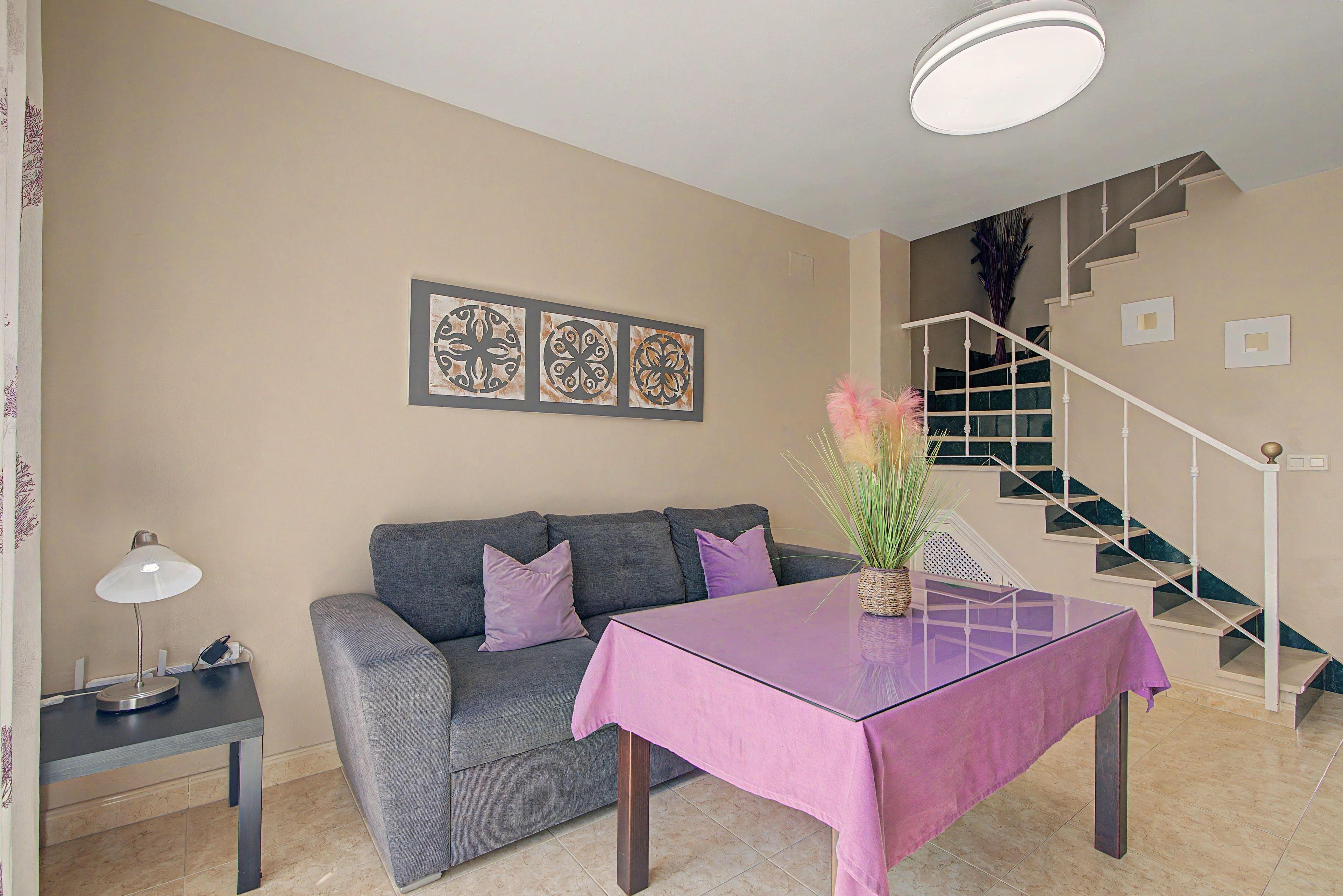 Una sala de estar con un sofá gris, una mesita con mantel rosa, una lámpara y una escalera con barandilla oscura.