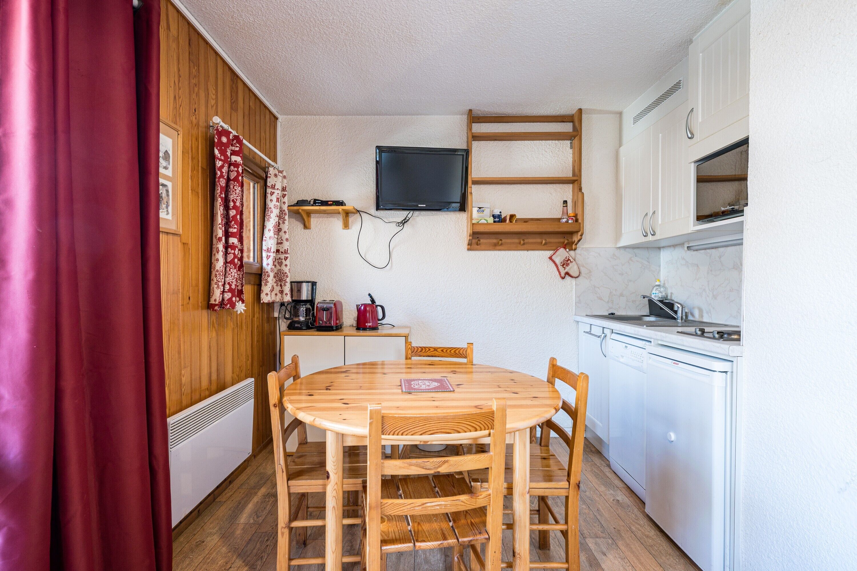 A compact kitchen with a round wooden table, white appliances, and a mounted TV.