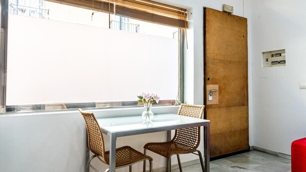 Una habitación con una mesa de vidrio, sillas de mimbre, un jarrón con flores, una puerta de madera y una ventana con persianas.