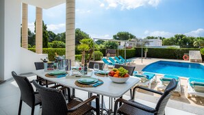 Zona de comedor junto a la piscina con una mesa para dos, un frutero y sillones de descanso alrededor de la alberca.