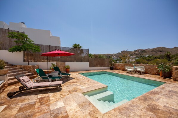 A swimming pool with lounge chairs and a red umbrella, surrounded by stone steps and potted plants.