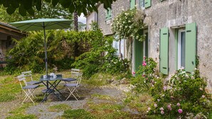 Een rustiek stenen gebouw met groene luiken, een tafel met stoelen en een tuin met bloeiende planten.