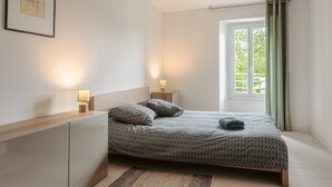 Een slaapkamer met een bed, nachtlampen, een nachtkastje, een raam met gordijnen en een foto in een lijst aan de muur.