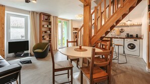 Een gezellige woonkamer met een eettafel, stoelen, een televisie en een bank. Een trap leidt naar een keukenruimte met een wasmachine.