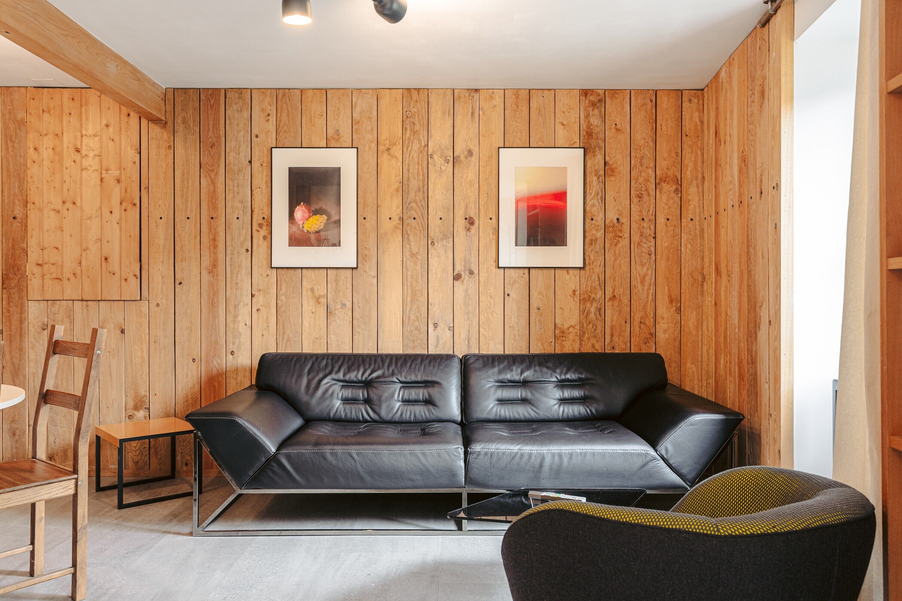 Een moderne woonkamer met een zwarte leren bank, een houten stoel, een klein tafeltje en twee ingelijste kunstwerken aan de houten wand.