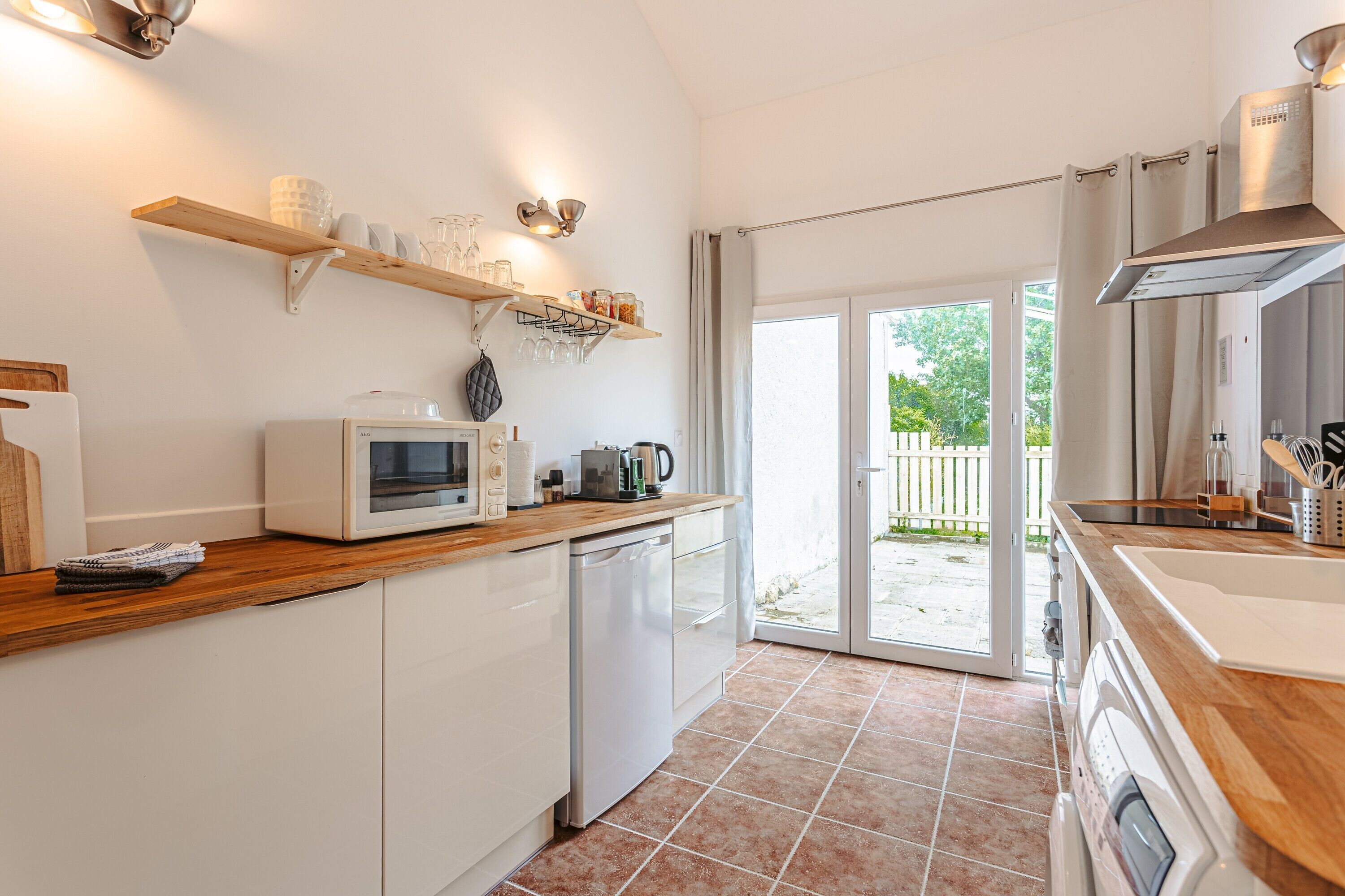 A modern kitchen with white cabinets, a microwave, a coffee maker, and a dishwasher. There is a wooden shelf above the microwave with glassware and jars. A sliding glass door leads to a patio with a wooden fence.