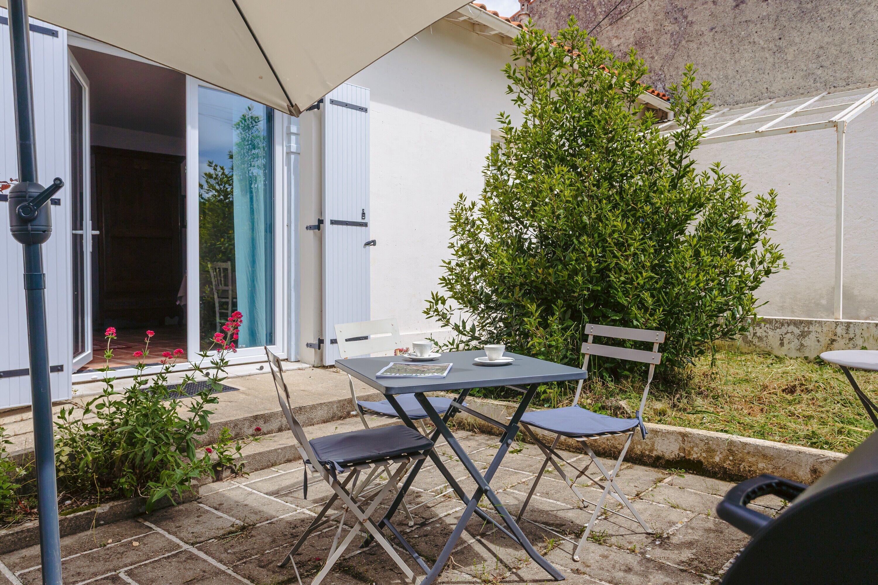 Een terras buiten met een tafel, stoelen, een kleine boom en een ingang van een gebouw.