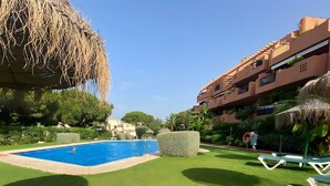 Ein von Grün umgebener Swimmingpool, ein Reetdachschirm und ein mehrstöckiges Gebäude mit Balkonen.