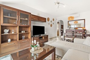 A living room with a TV, wooden cabinets, a glass coffee table, and a dining area with chairs and a table.