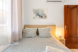 A bedroom with a bed, a nightstand with a lamp, a wooden door, and a wall-mounted air conditioner.