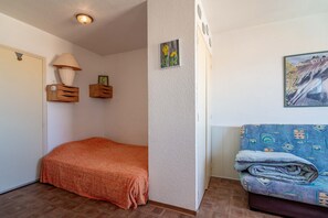 Un dormitorio con cama, mesita de noche, lámpara, estante en la pared con un cuadro y sofá con manta.