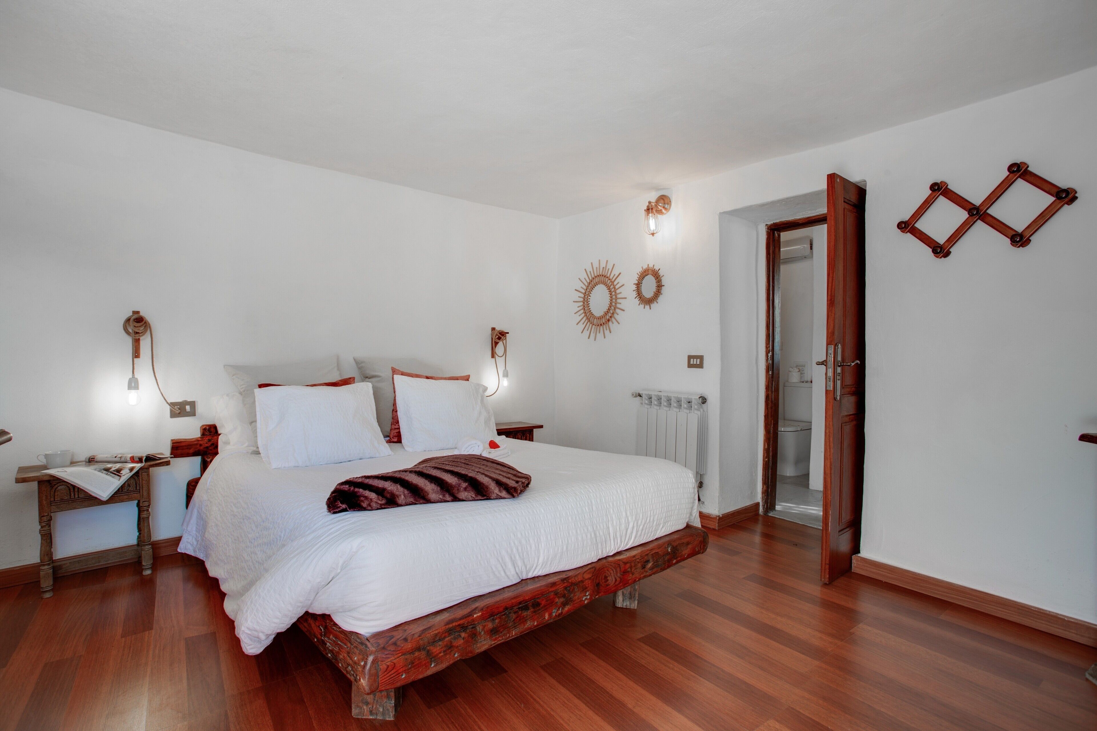Une chambre à coucher avec un lit en bois, du linge de lit blanc, une table de chevet, une lampe de lecture et une porte donnant sur une salle de bain.