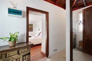 Ein Zimmer mit einem Holzkasten, einer Topfpflanze, einem Bett mit weißer Bettwäsche und einer Tür zu einem anderen Raum.