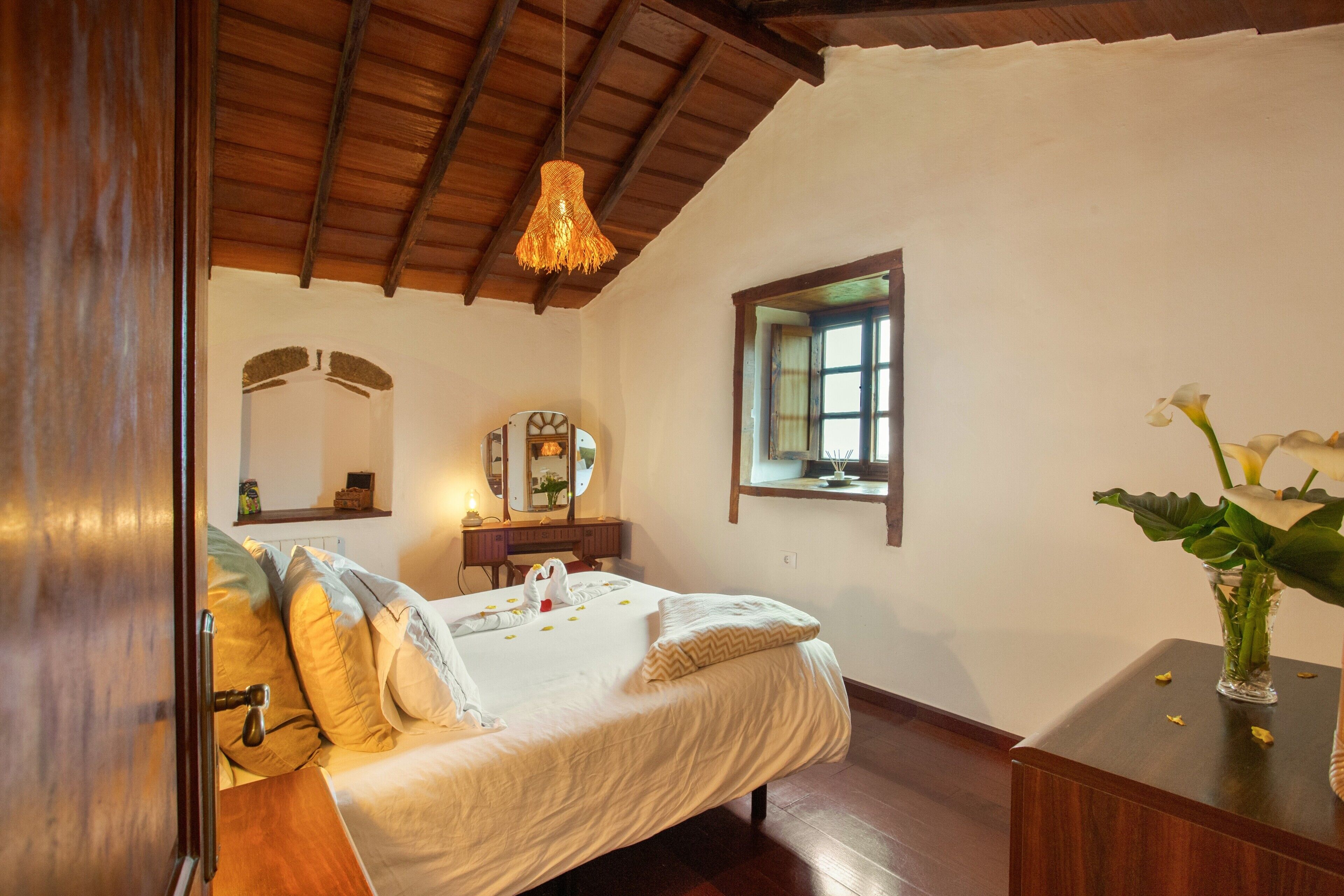 A bedroom with a wooden ceiling, a bed with white bedding, a bedside table with a vase, and a window with a view.