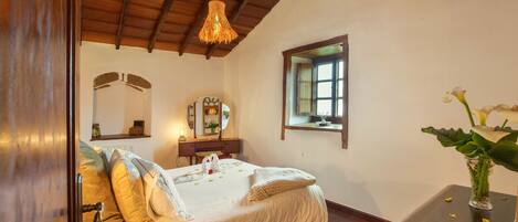 A bedroom with a wooden ceiling, a bed with white bedding, a bedside table with a vase, and a window with a view.