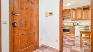 A wooden door with a lock, a kitchen with wooden cabinets, a tiled floor, and a small table with a chair.