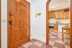 Una puerta de madera con cerradura, una cocina con armarios de madera, suelo de baldosas y una pequeña mesa con una silla.