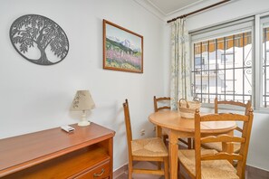 A dining area with a round wooden table, four chairs, a lamp, a framed picture, and a window with metal bars.