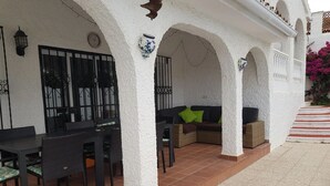 Eine überdachte Terrasse mit Esstisch und Stühlen, einer Couch mit Kissen sowie einer Wand mit einer Dekoration und einer Uhr.