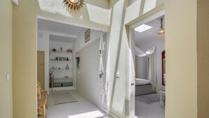 Un couloir avec un mur blanc, une chaise en bois, une étagère avec des livres et des plantes, et un lit à l’arrière-plan.