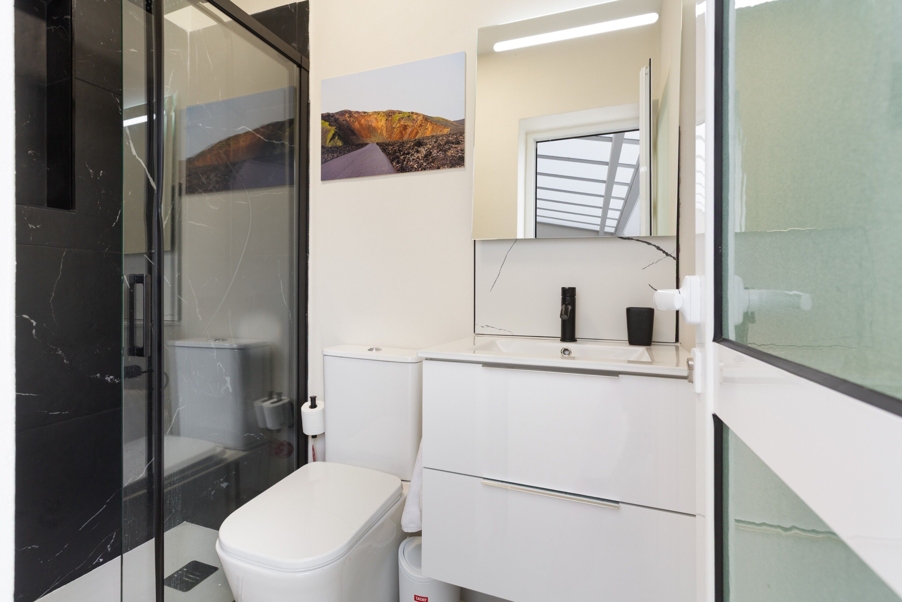 Ein modernes Badezimmer mit Duschkabine aus Glas, einer weißen Toilette, einem weißen Waschbecken mit Spiegel und einem Landschaftsbild an der Wand.
