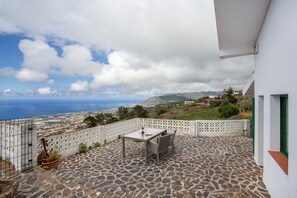 Eine Steinterrasse mit Tisch und Stühlen, ein weißer Zaun und Blick auf das Meer und die Hügel.
