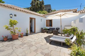 Un patio con un edificio blanco, suelo de piedra, plantas en macetas, una silla de descanso y una mesa.