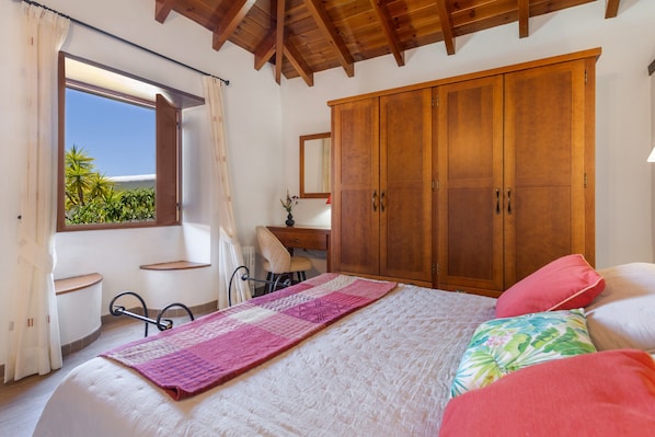 A bedroom with a bed, a wooden wardrobe, a desk, and a window with a view of greenery.