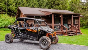 Un véhicule tout-terrain Polaris RZR est garé devant une cabane rustique en rondins de bois, dotée d’une véranda.
