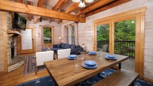A wooden cabin interior with a dining table set for four, a fireplace, a sofa, and large windows offering a view of the outdoors.