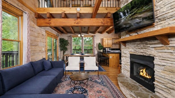 A log cabin interior with a sofa, dining table, chairs, a fireplace, and a TV mounted on a stone wall.