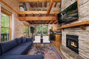 A log cabin interior with a sofa, dining table, chairs, a fireplace, and a TV mounted on a stone wall.