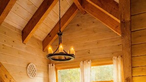 A cozy bedroom with a wooden ceiling, a bed with white bedding, two pillows, a hanging light fixture, and a window with curtains.