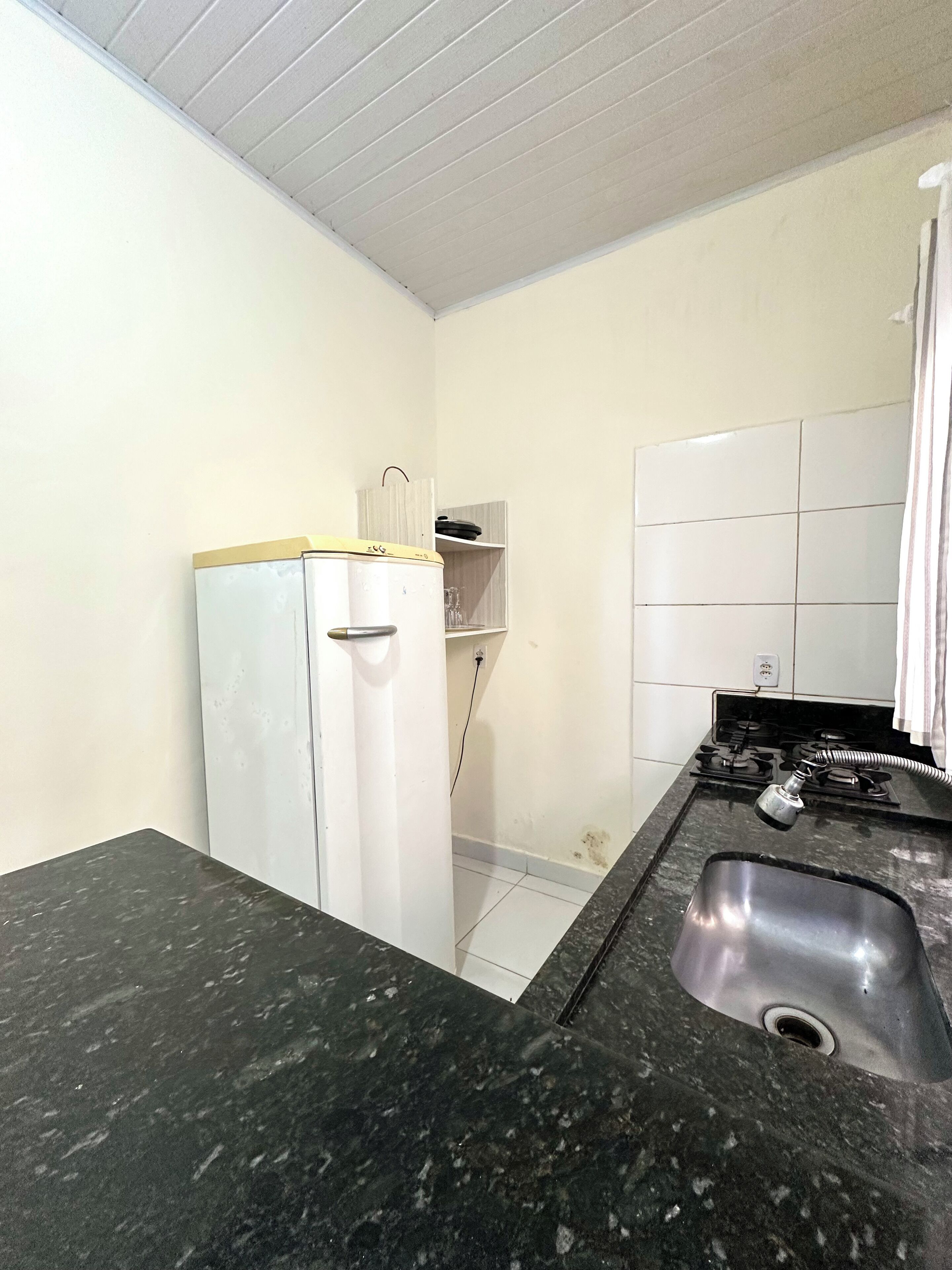Uma cozinha compacta com bancada de granito preto, geladeira branca, fogão a gás e uma pequena estante com uma chaleira.
