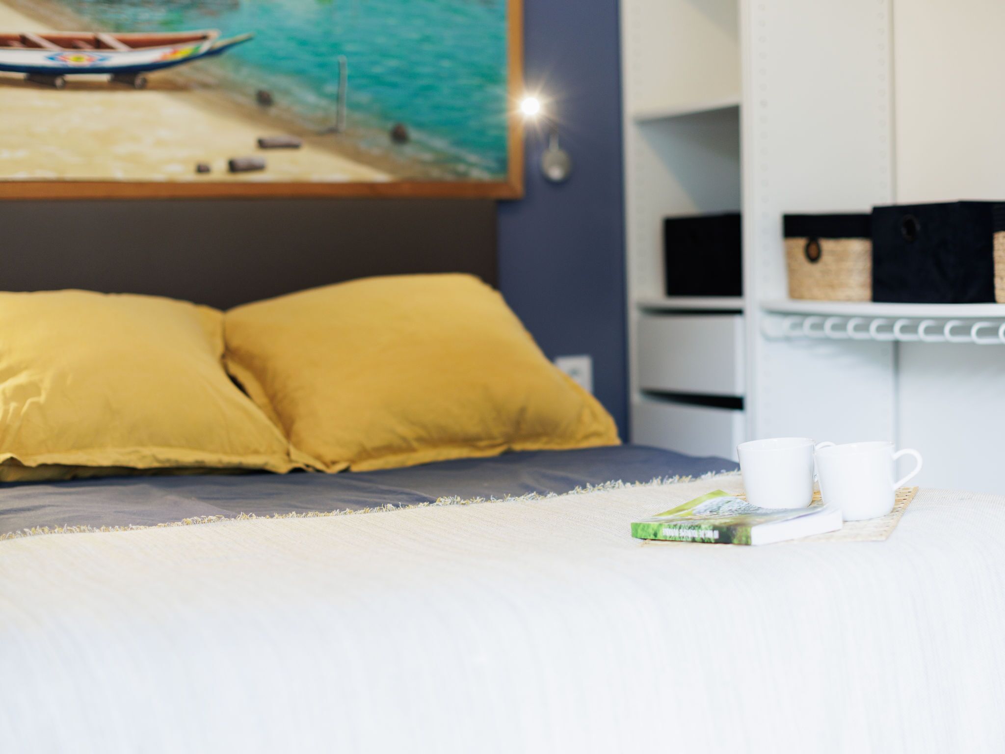 A bed with two yellow pillows, a white blanket, and two white cups on a shelf.