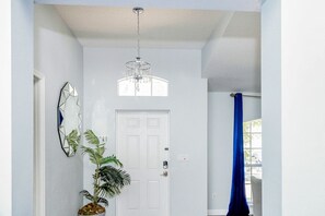 Stylish hallway with a simple door, geometric mirror, and potted plant, leading to a room with a chandelier.