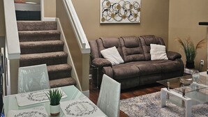 A modern living room with a brown leather sofa, a glass coffee table, and a dining area with a table set for two. A staircase with a white railing leads to an upper level. - Charming 3-bedroom house in awesome Urbandale! (Urbandale)