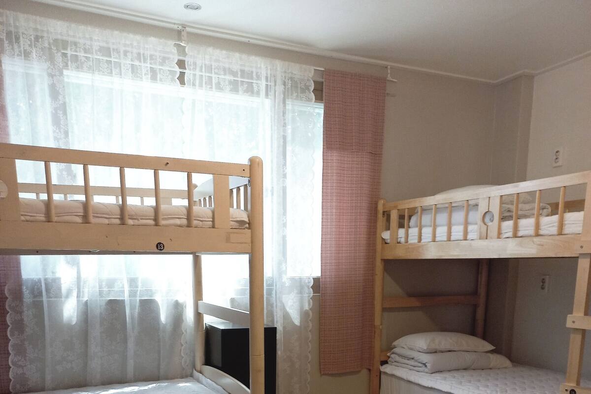 A room with two bunk beds, wooden flooring, a window with sheer curtains, and a small bedside table.