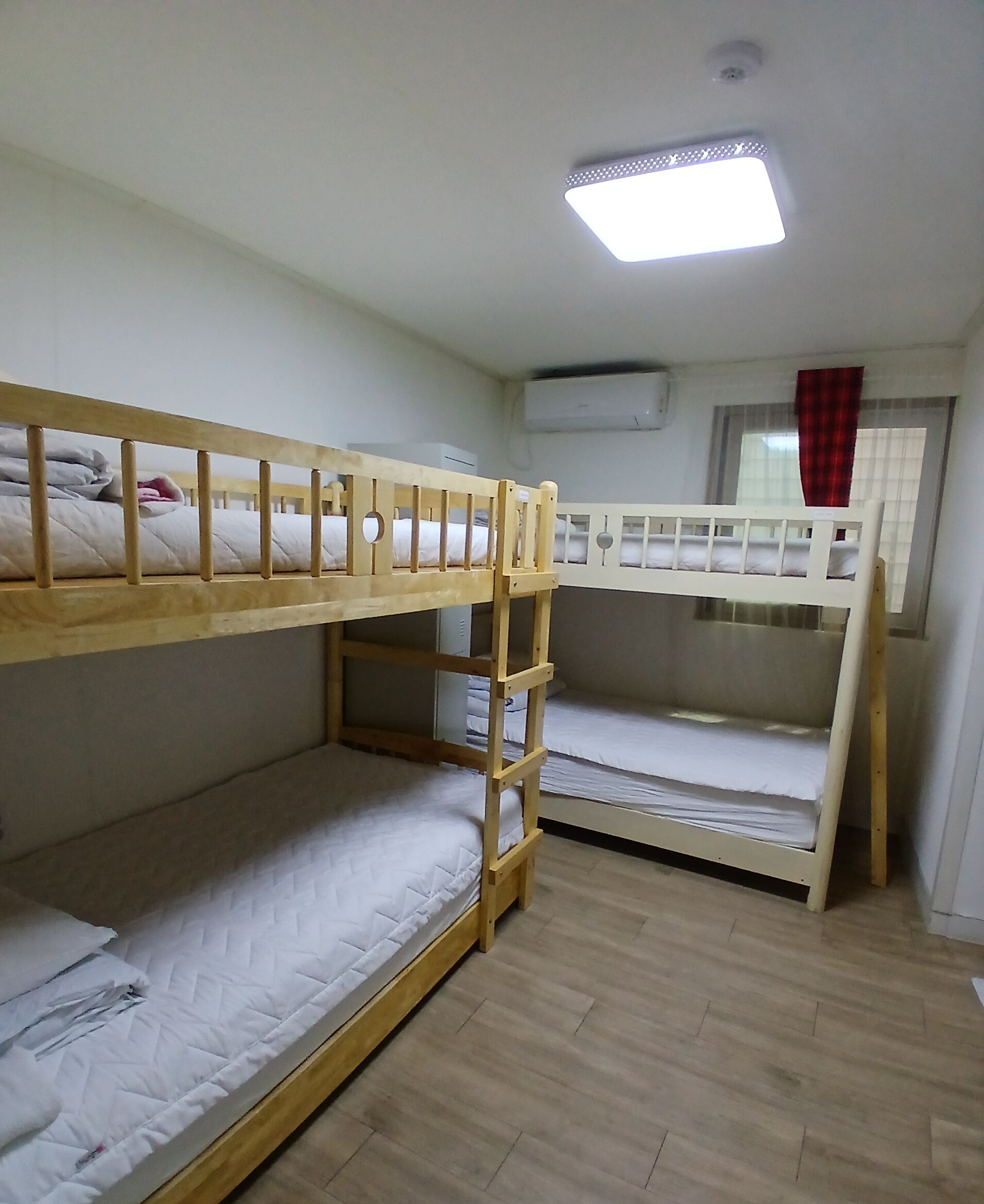 A dormitory room with bunk beds, a ceiling light, an air conditioning unit, and a window with curtains.
