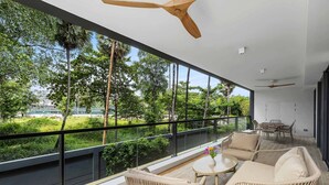 Un balcón con mobiliario de mimbre, un ventilador de techo y vistas a la vegetación y un edificio a lo lejos.