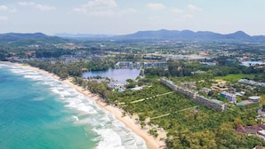 Vista aérea de un complejo turístico costero con propiedades frente a la playa, un lago y montañas al fondo.