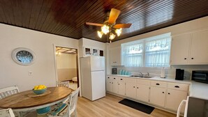 Una cocina con armarios blancos, techo de madera, ventilador de techo, un reloj, una mesa de comedor con sillas y una ventana con cortinas.
