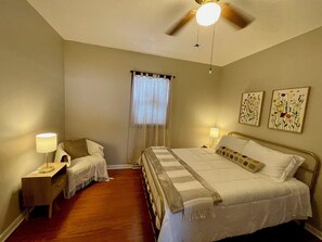 A bedroom with a bed, a nightstand, a lamp, a ceiling fan, a window with curtains, and two wall art pieces. - The Little House on Prairie: Family-Friendly 2-Bedroom Getaway (Mattoon)