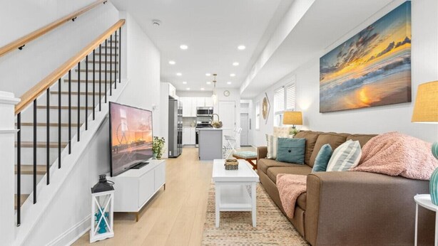 A modern living room with a staircase, a sofa, a coffee table, and a painting of a sunset. - Modern 3BR w/ Backyard, Grill & Outdoor Shower (Ventnor City)